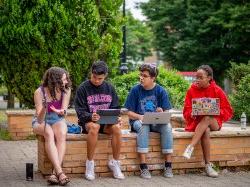 Four students sitting on brick ledge outside of Dickson Hall during the Summer Bridge Program researching different campus resources on laptops and notebooks.